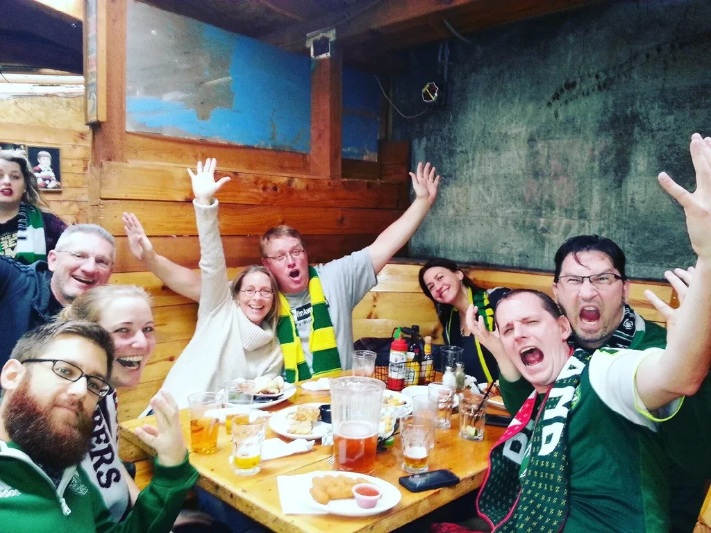 Eight people in a bar cheer, wearing green and yellow scarves.