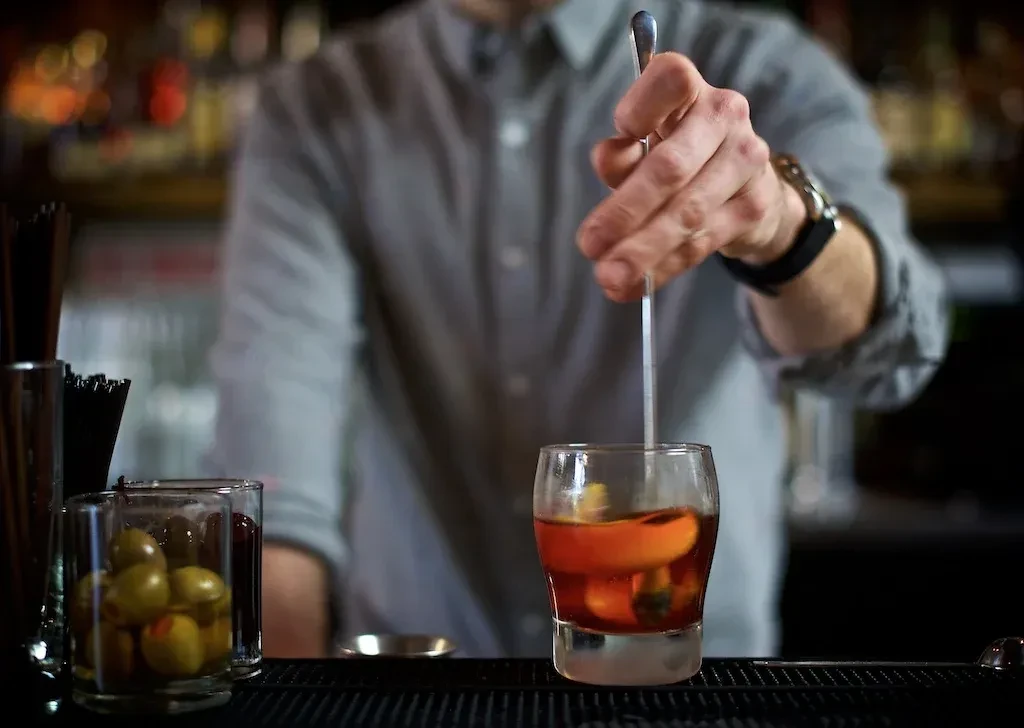 Bartender's hands stirring a cocktail with orange peel and olives.