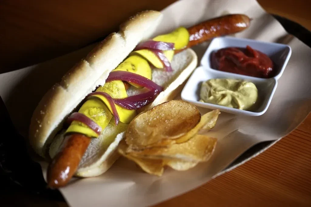 Gourmet hot dog with bright pickles, red onions, chips, ketchup, mustard.