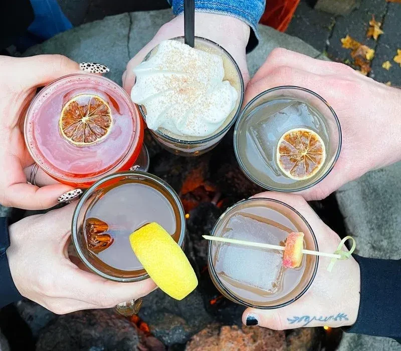 Overhead view of five hands holding various garnished drinks near a fire.