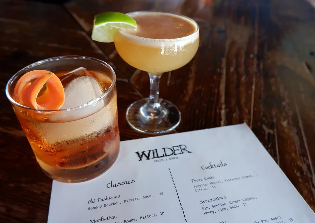 Two cocktails on a dark wooden table with orange peel and lime.