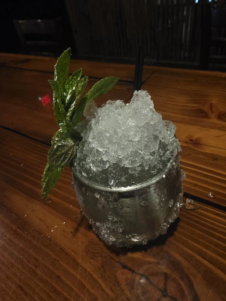 Overflowing silver metal cup with crushed ice, mint, and straw on wood.