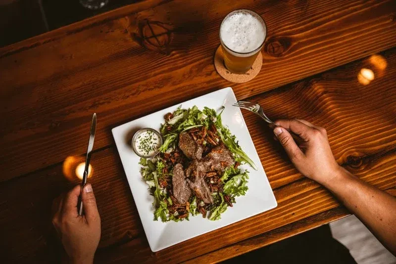 Hands cutting a grilled meat and pecan salad with beer.