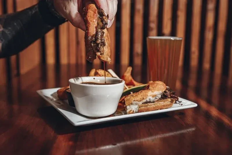 Tattooed hand dips cheesesteak into sauce, with fries and beer.