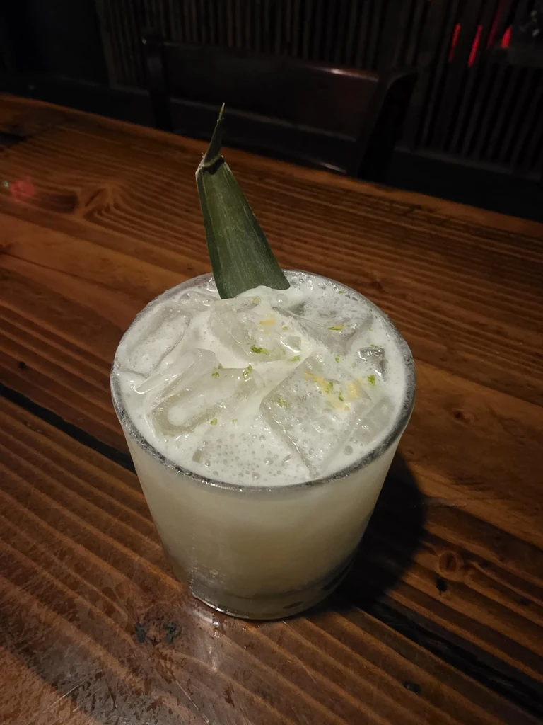 Close-up: Foamy, light cocktail with ice, green specks, and leaf garnish.