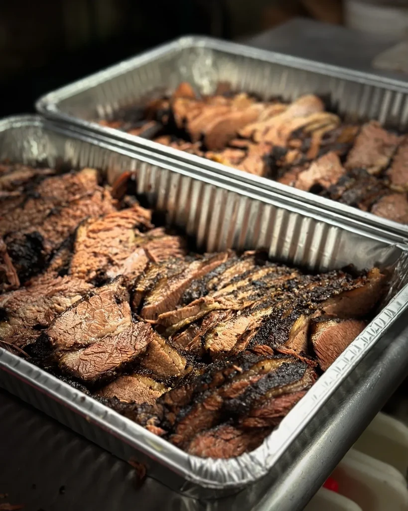 Two aluminum trays of sliced barbecue brisket with dark bark.