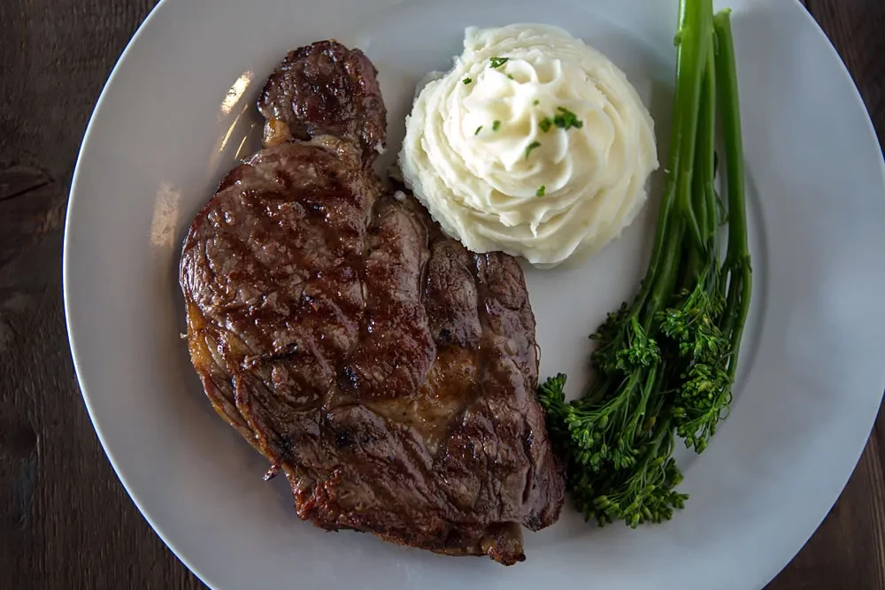 Grilled steak, mashed potatoes, and broccolini on a white plate.