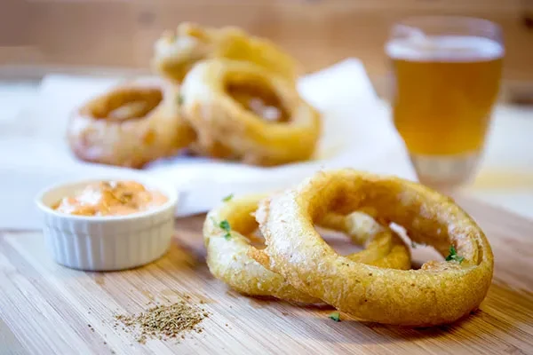 Golden-brown onion rings, orange dipping sauce, and herbs on a wooden board.