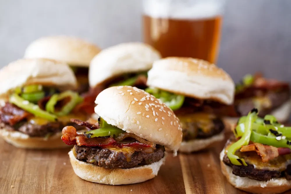Miniature burgers with cheese, bacon, peppers, and beer on wood.