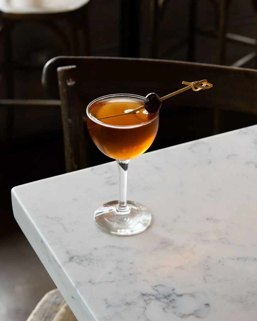 Amber cocktail in coupe glass with cherry garnish on marble table.