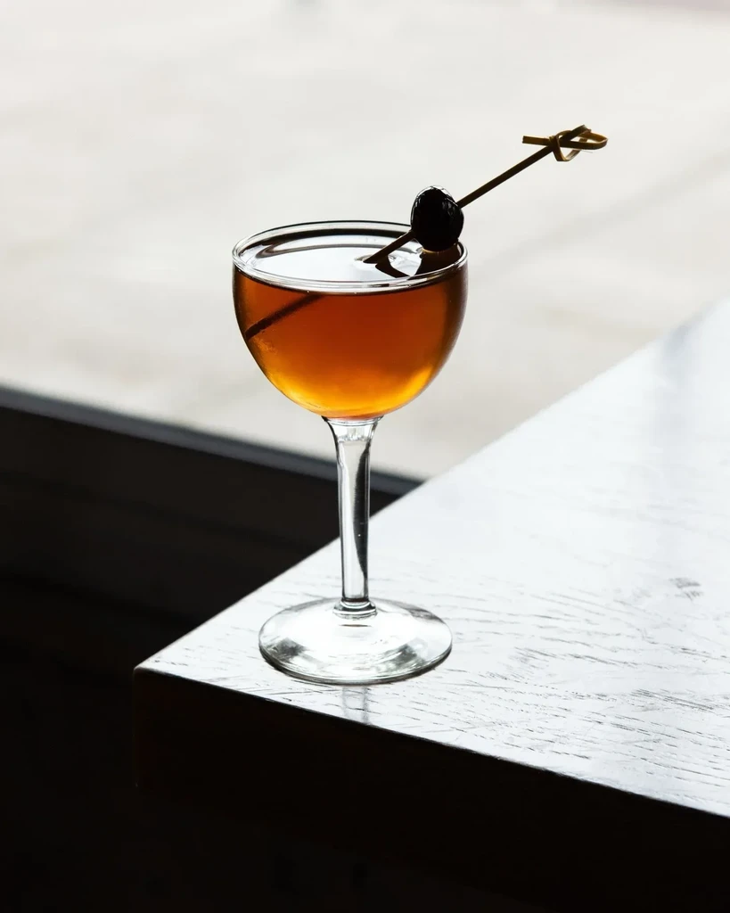 Amber cocktail in coupe glass with cherry on a wooden table.