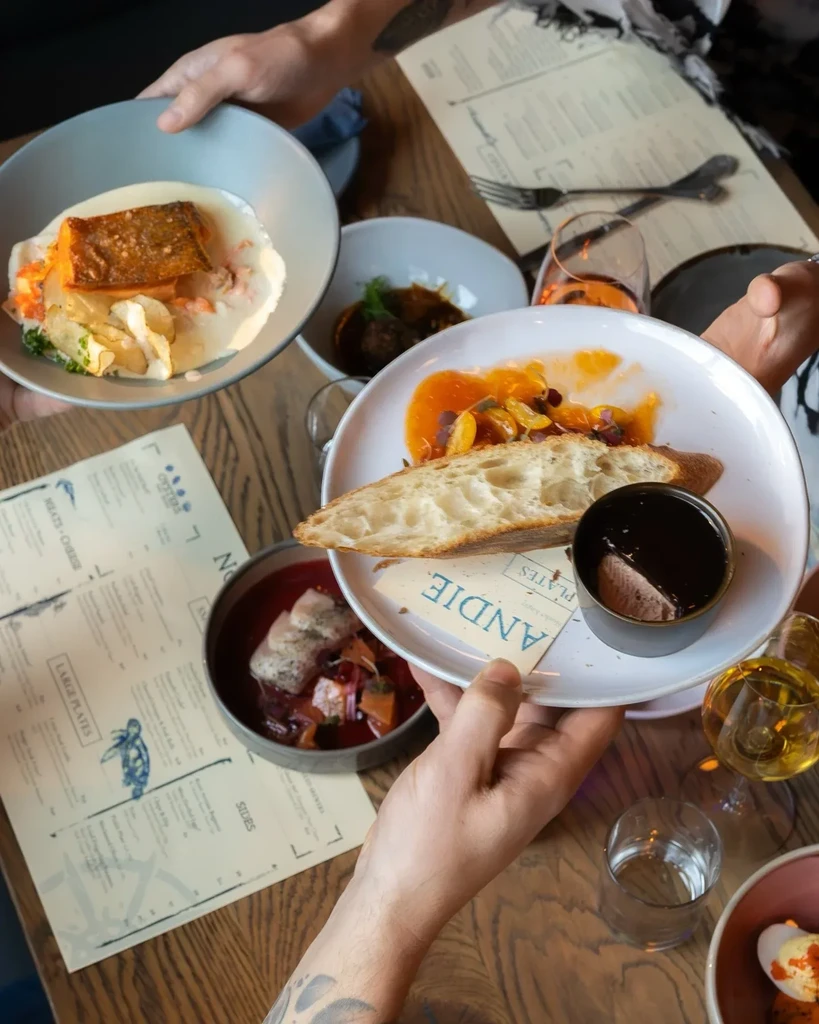 Overhead view of a restaurant table with diverse food, drinks, and hands.