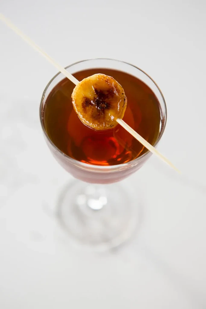 Amber cocktail in stemmed glass, garnished with skewered caramelized banana slice.