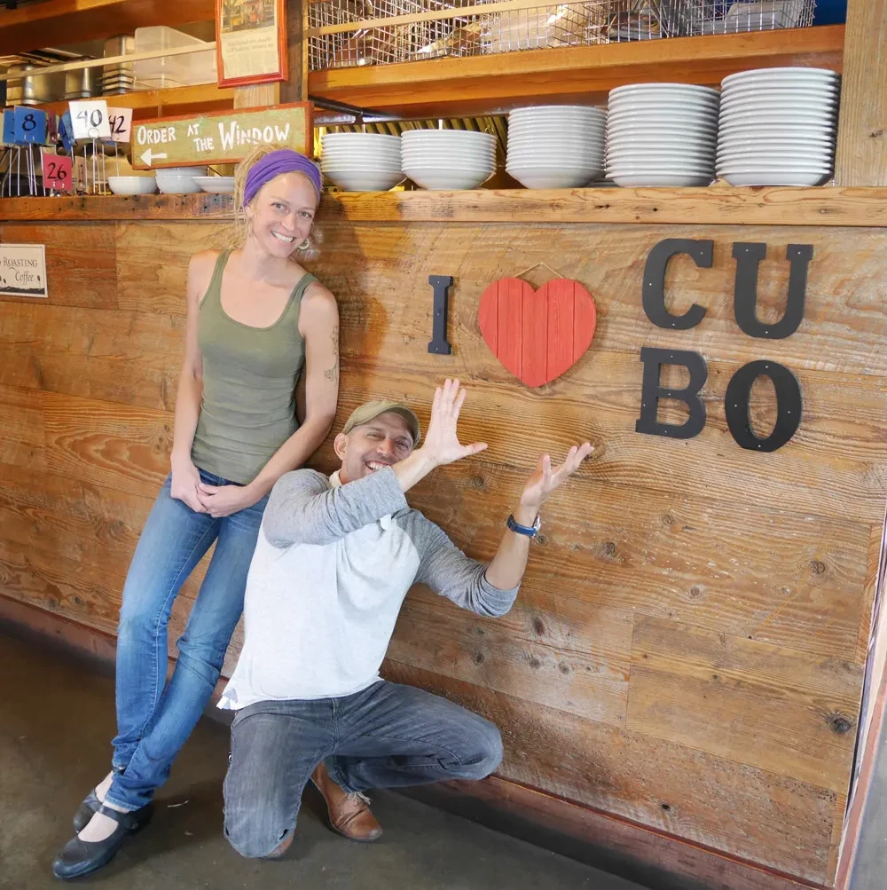 Man and woman smiling in a rustic cafe, man making 'L' gesture.