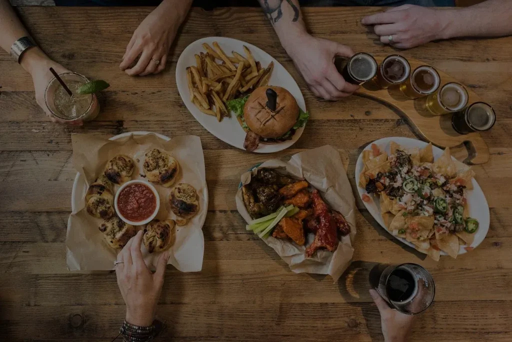 Rustic table laden with burgers, nachos, wings, beers, and hands.