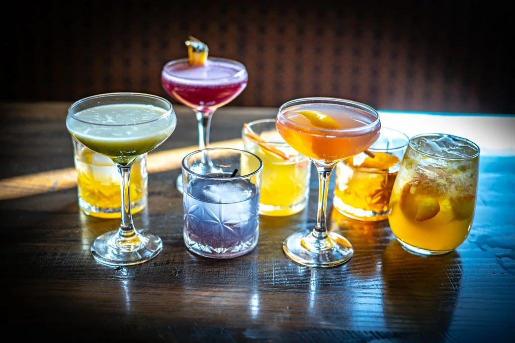 Seven vibrant, garnished cocktails in various glasses on a dark bar.