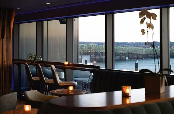 Modern restaurant interior overlooks river and green truss bridge at dusk.