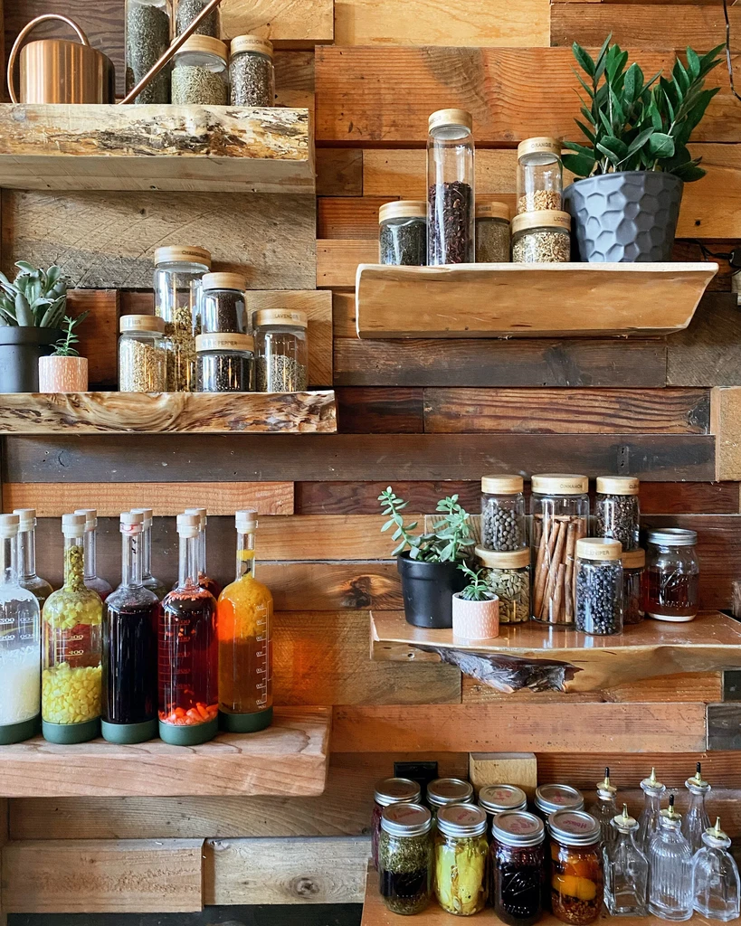 Jars of spices, infused liquids, and plants on rustic wooden shelves.