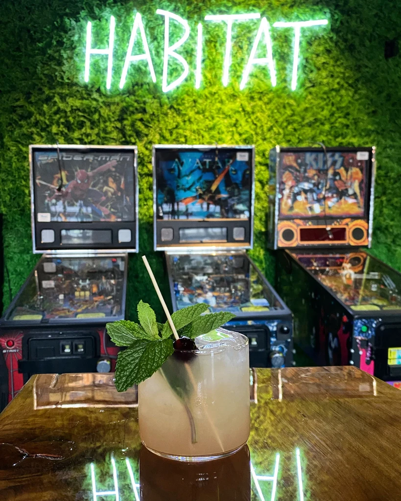 Pale cocktail on bar, 'HABITAT' neon, pinball machines, and moss wall.