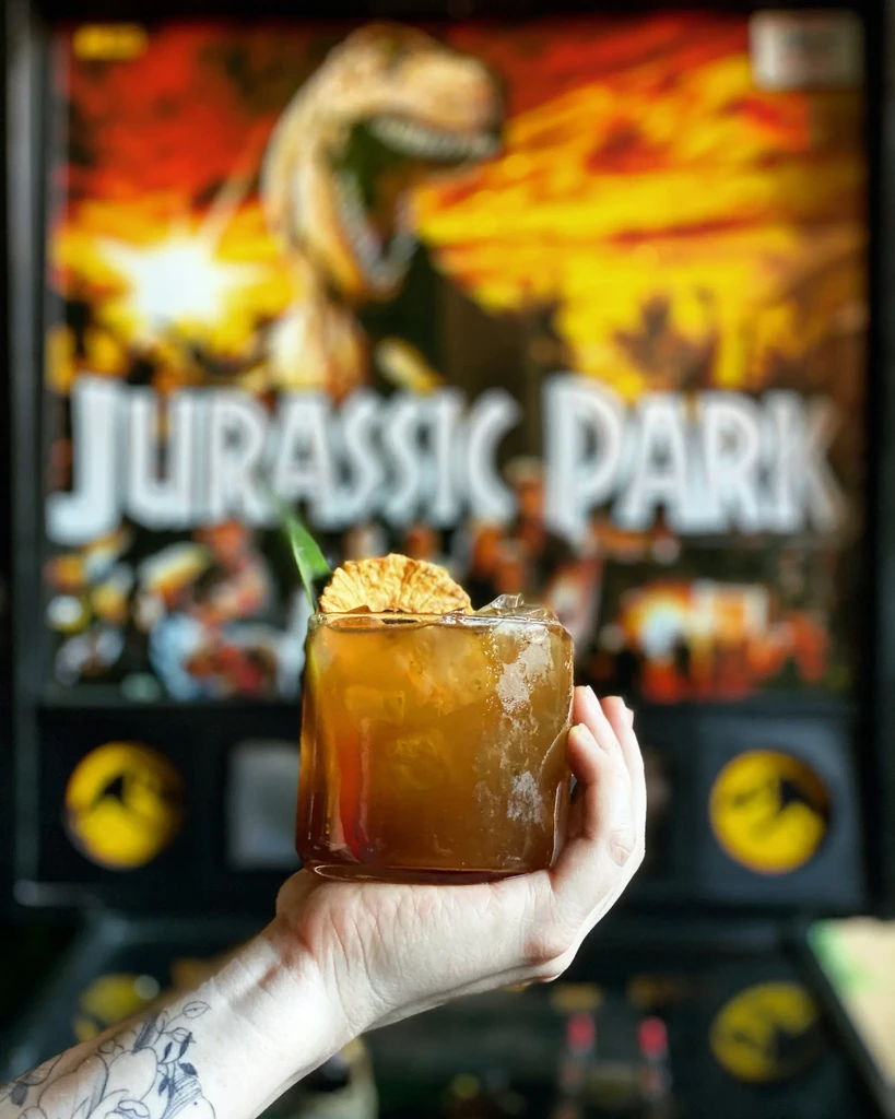 Tattooed hand holds garnished amber cocktail, Jurassic Park pinball background.