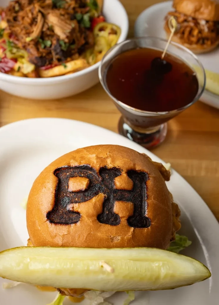 PH-branded burger with pickle, cocktail, pulled meat, and slider on table.