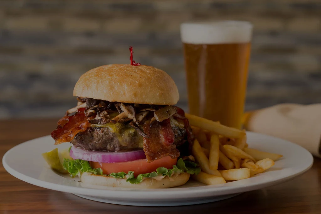 Gourmet burger with melted cheese, bacon, fries, and beer on a table.