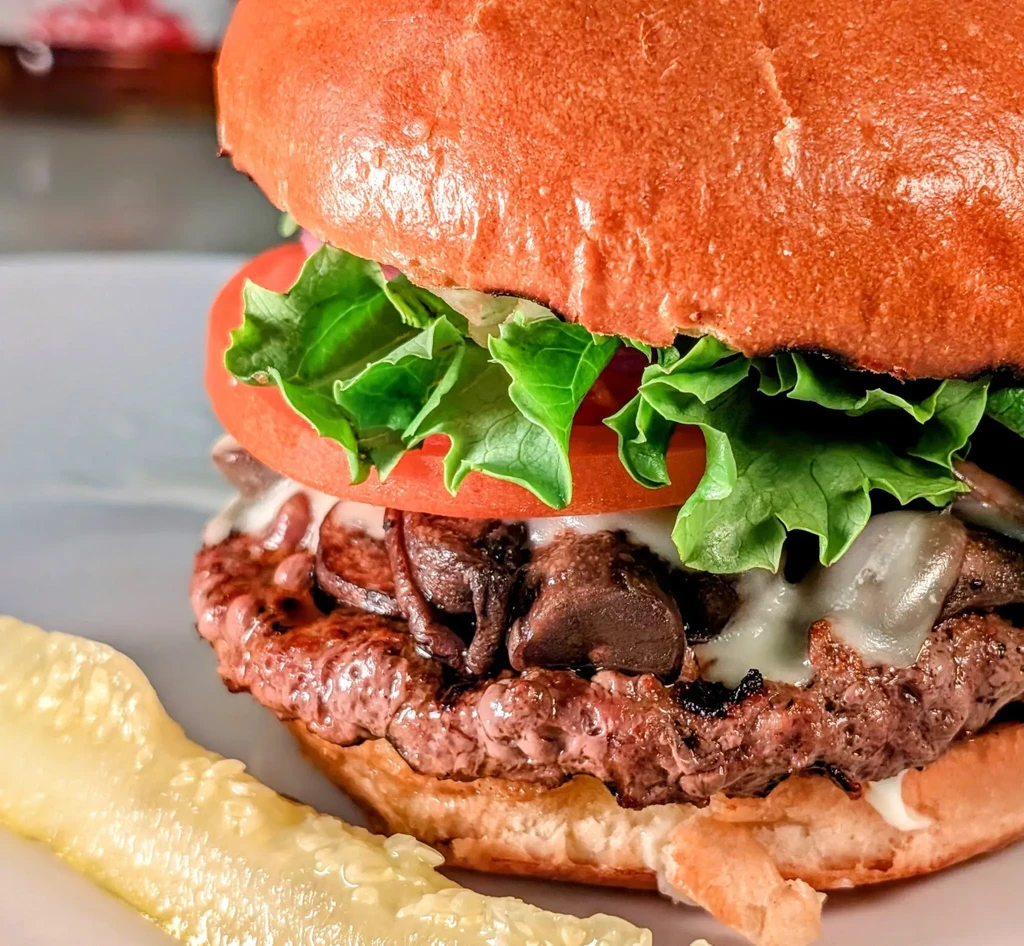 Gourmet beef burger with lettuce, tomato, cheese, mushrooms, and a pickle.