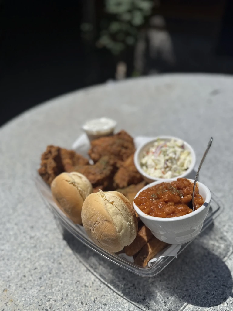 Fried chicken, coleslaw, baked beans, rolls in takeout container outdoors.