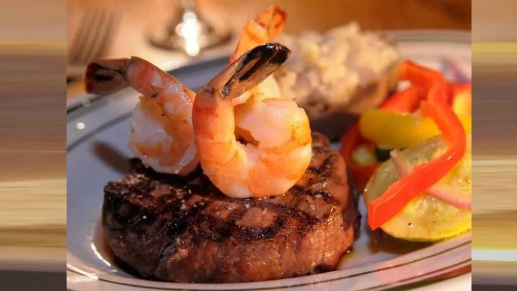 Grilled steak topped with two shrimp, vibrant vegetables, and a creamy side.