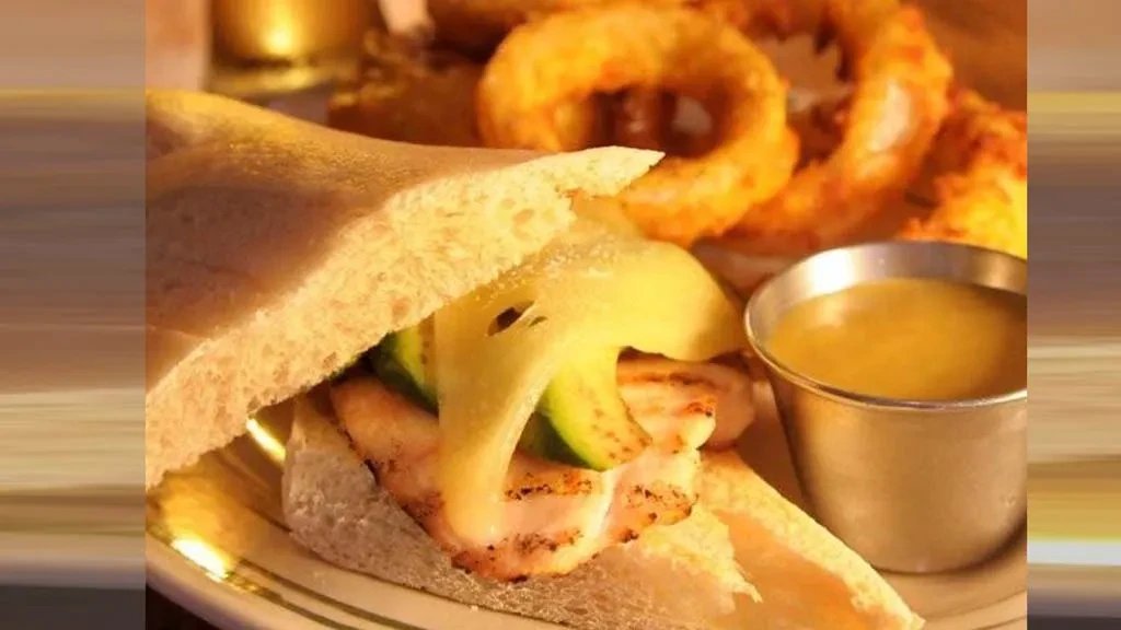 Large grilled chicken sandwich, melted cheese, green vegetables, sauce, and onion rings.