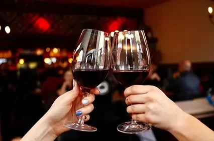 Two hands clink red wine glasses in a blurred restaurant.
