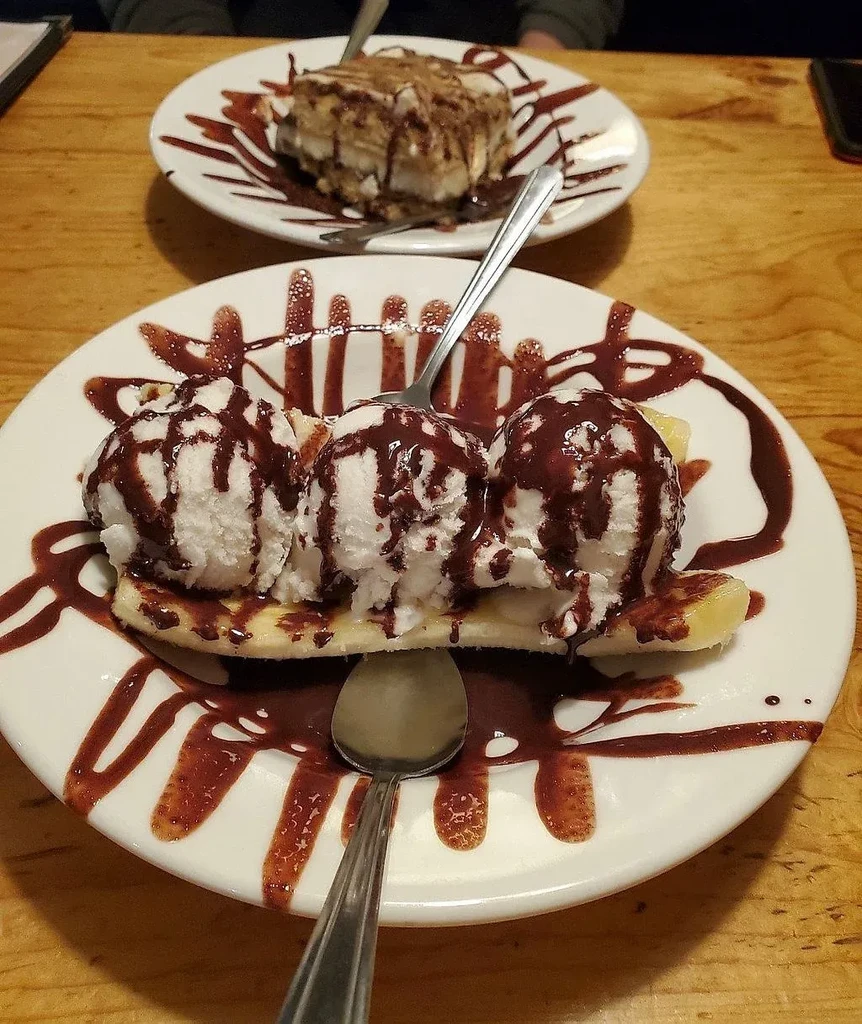 Chocolate-drizzled banana split and blurred cake on a wooden table.
