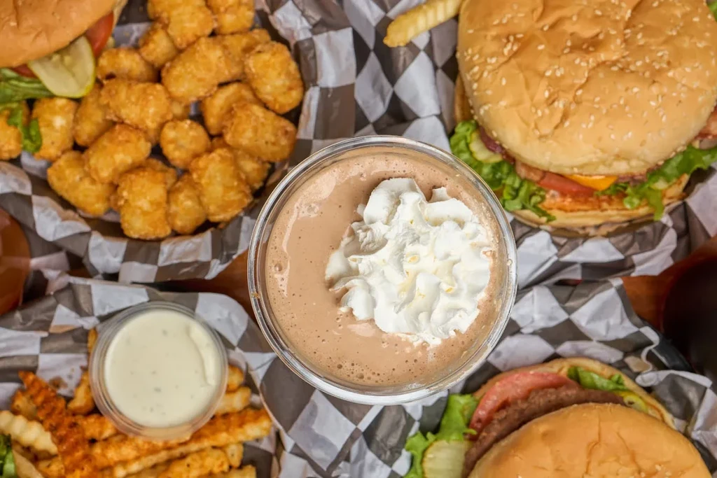 Overhead view of a chocolate milkshake surrounded by burgers, fries, and tots.