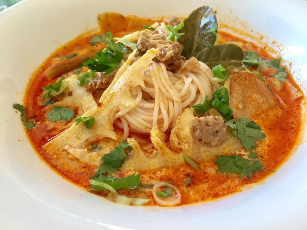 Overhead: creamy reddish-orange noodle soup with meat, thin noodles, herbs.