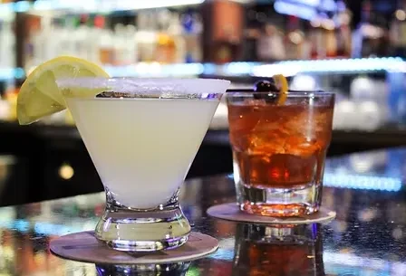 Two cocktails: light with lemon, dark with cherry, on a bar.