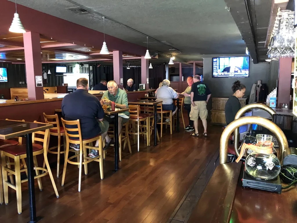 Casual restaurant interior with patrons, high-top tables, bar, and a TV.