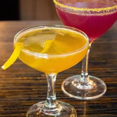 Two cocktails: yellow coupe with lemon, purple martini with sparkly rim.