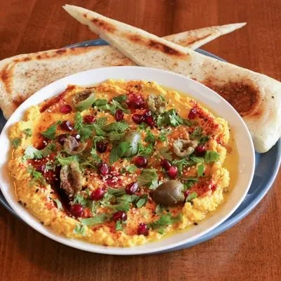 Yellow hummus with herbs, pomegranate, olives, and flatbread on wood.