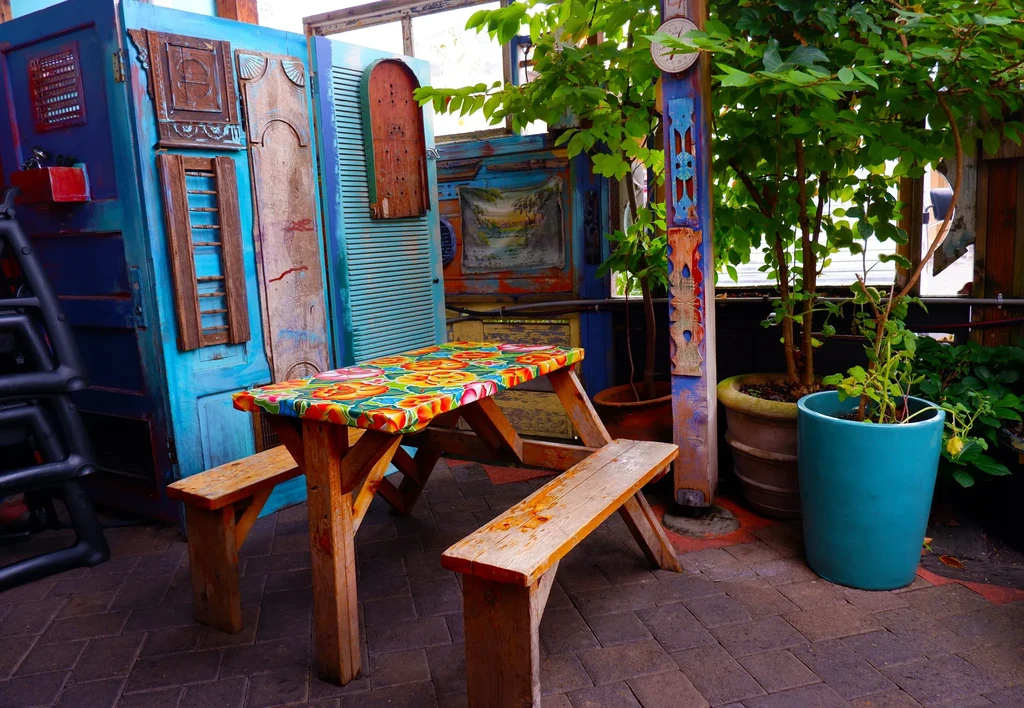 Rustic picnic table, floral cloth, blue patchwork wall, and potted plants.