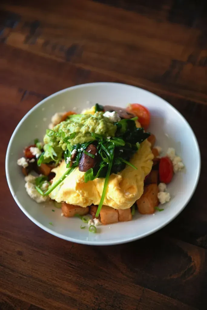 Breakfast bowl with scrambled eggs, guacamole, potatoes, spinach, mushrooms, tomatoes, cheese.