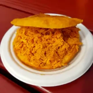 Close-up: open-faced arepa with shredded chicken on a paper plate.