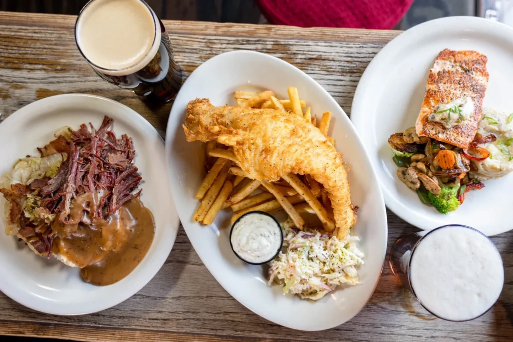 Rustic table laden with fish and chips, salmon, shredded meat, and beer.