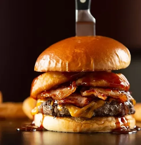 Gourmet burger with beef, melted cheese, bacon, onion rings, held by knife.