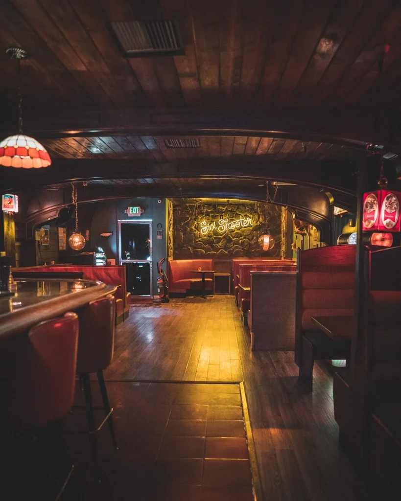Cozy bar interior with red seating, wooden ceiling, "Six Shooter" neon sign.