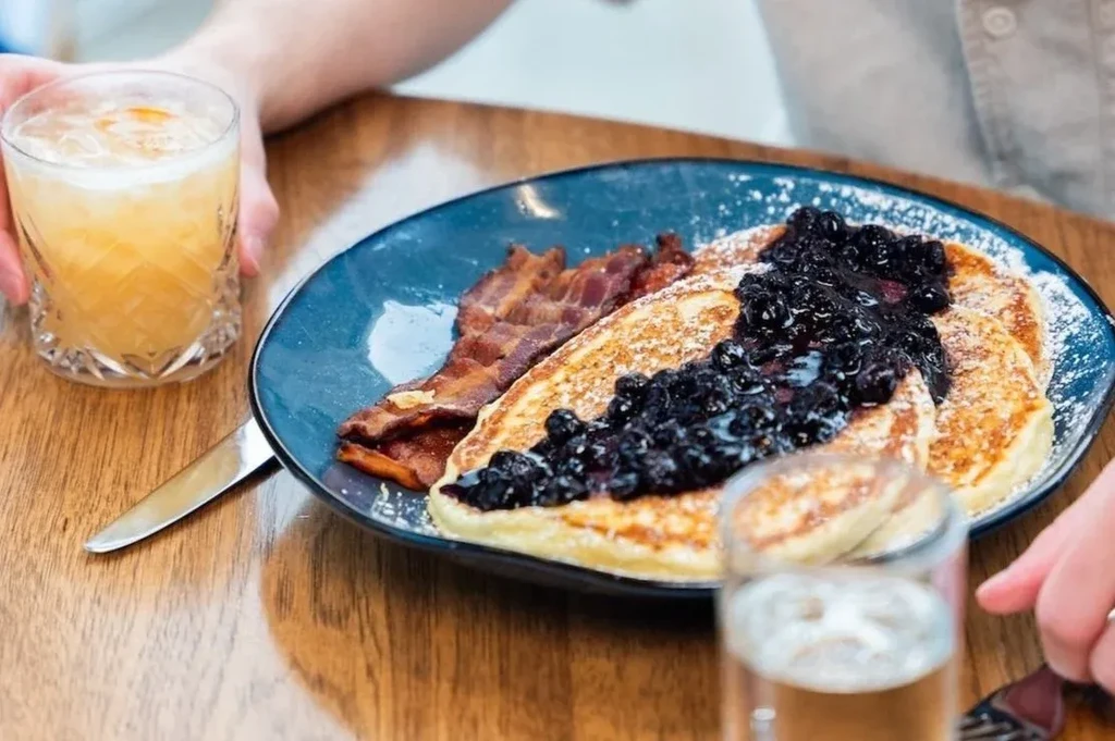 Pancakes with blueberry compote, bacon, and amber drink on table.