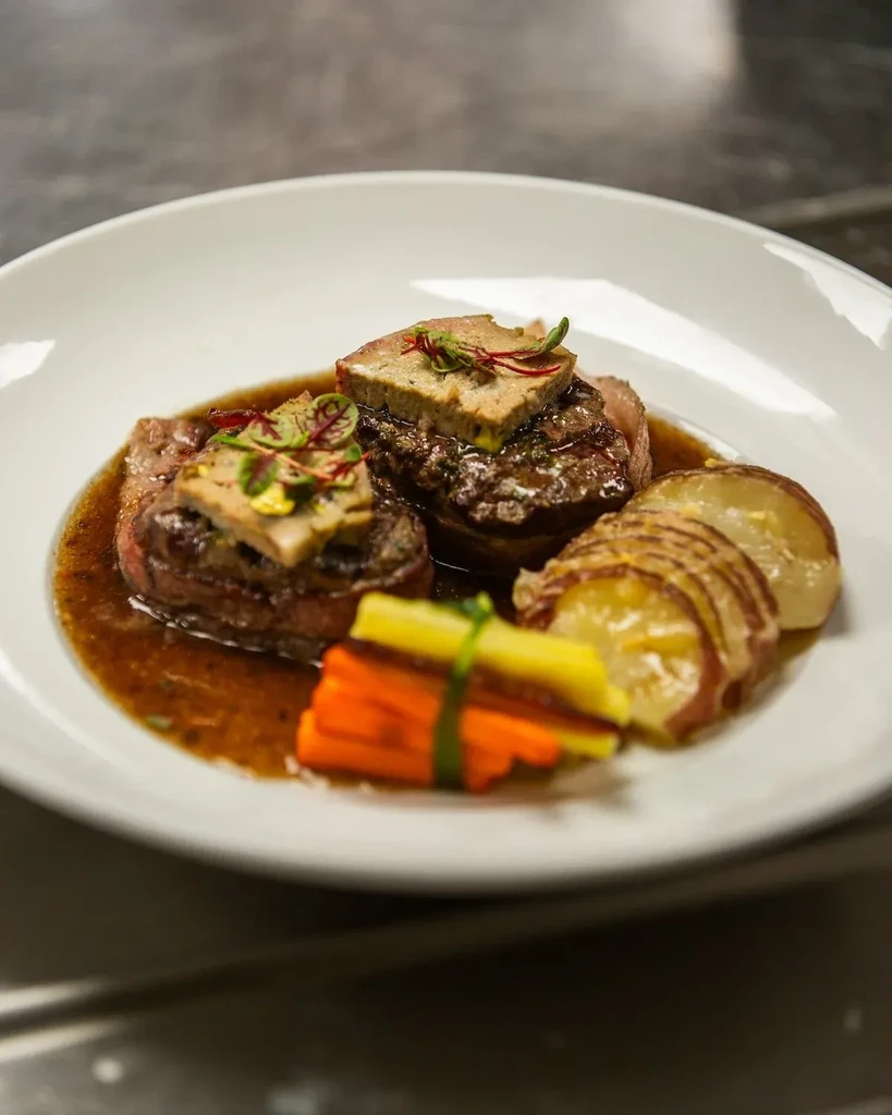 Plated beef medallions topped with pâté, sliced potatoes, and tied vegetables.