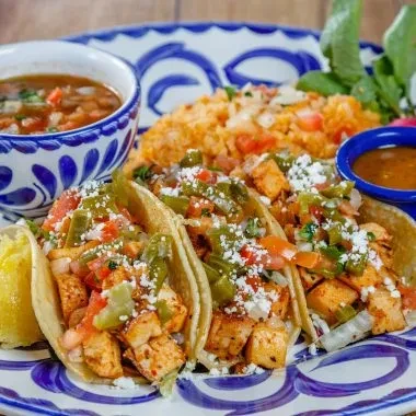 Three chicken tacos, Mexican rice, soup, and sauce on a blue plate.