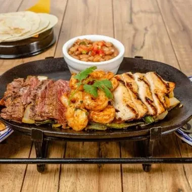 Sizzling platter with grilled steak, chicken, shrimp, beans, and tortillas.