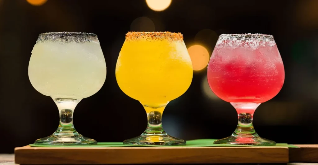 Three frosted, colorful cocktails with unique rims on a wooden board.