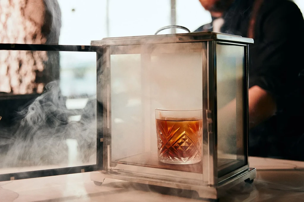 Amber cocktail in glass smoking box, smoke billowing inside.